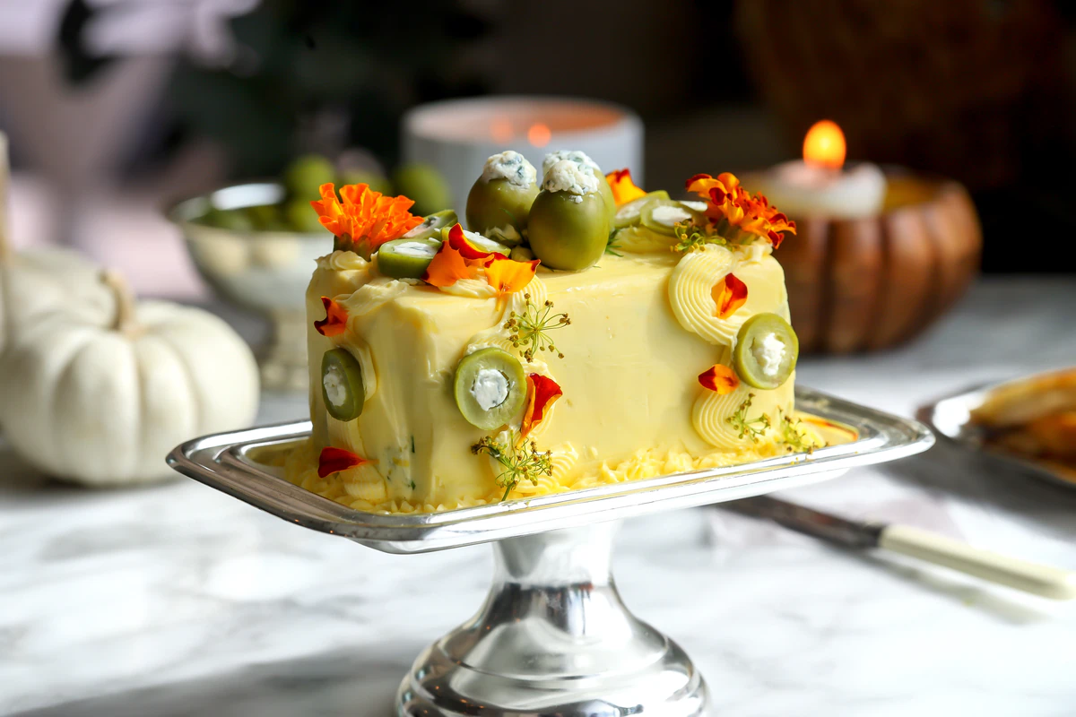 A dirty martini butter cake on a silver pedestal stand topped with blue cheese-stuffed olives, orange edible flowers, and dill flowers
