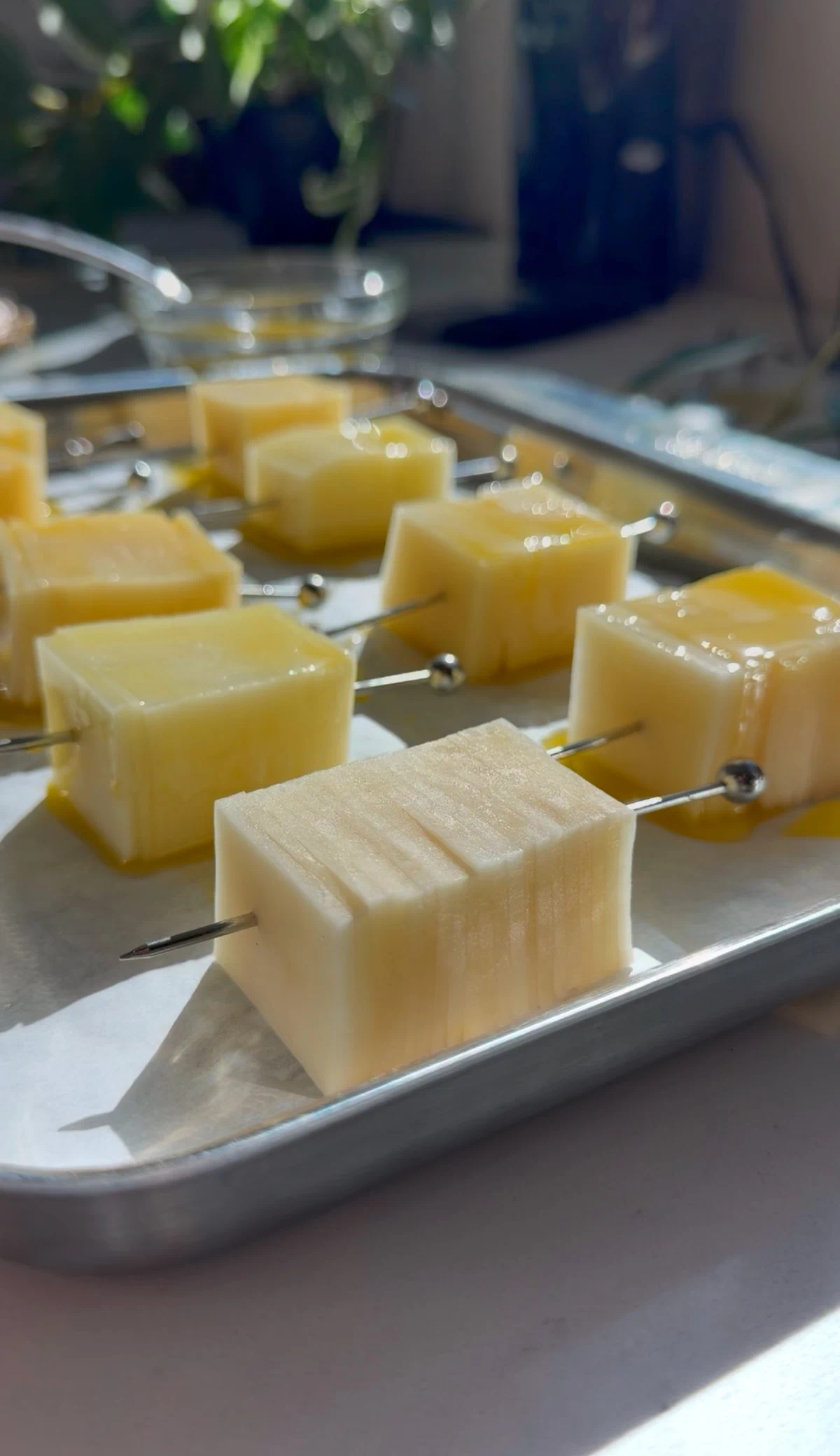 Stacks of thinly sliced raw potatoes on metal cocktail picks, brushed with clarified butter on a parchment-lined baking sheet.