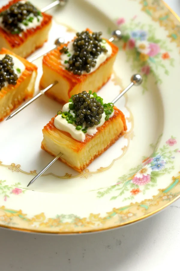 Domino potato skewers topped with crème fraîche, chives, and caviar on a vintage floral china plate, showing the crispy golden layers.