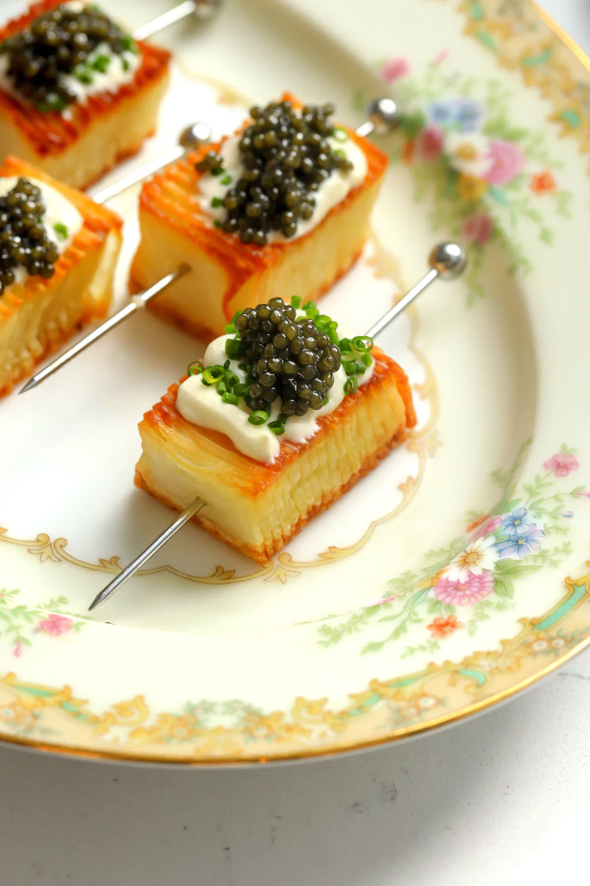 Domino potato skewers topped with crème fraîche, chives, and caviar on a vintage floral china plate, showing the crispy golden layers.