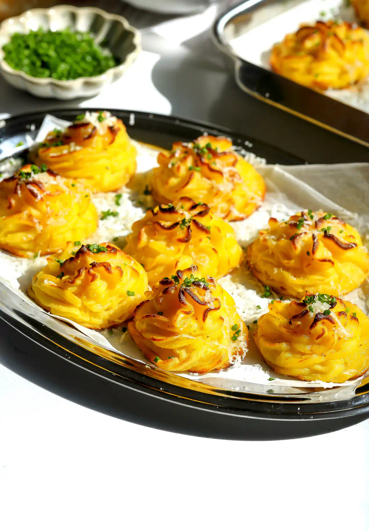 Gruyère stuffed duchess potatoes with browned piped ridges, Parmesan, and chives on parchment in an oval tray, with a bowl of chives behind.