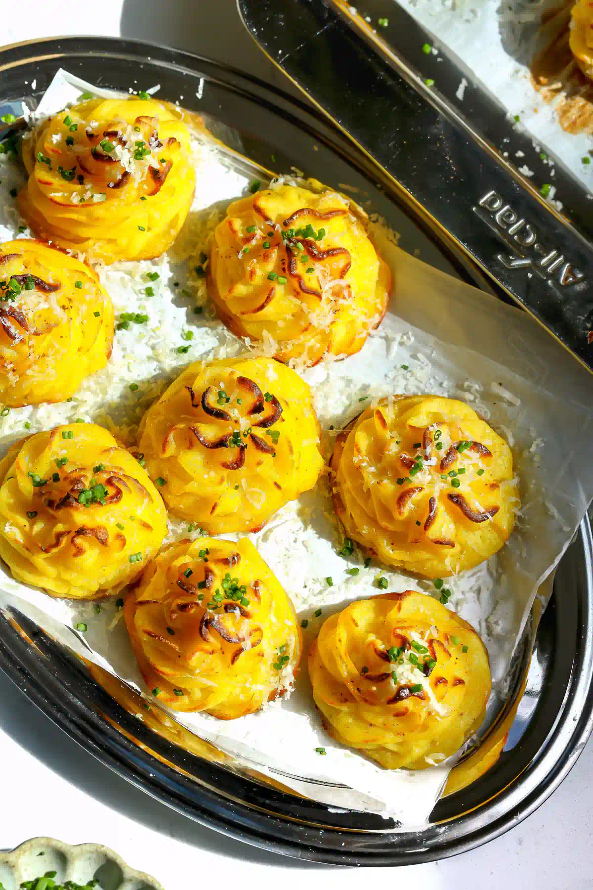 Overhead view of eight duchess potatoes topped with grated Parmesan and chives on parchment in a dark oval tray, next to a sheet pan.