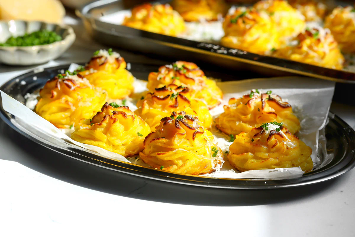Golden baked duchess potatoes with Parmesan and chives on parchment in an oval tray, with a sheet pan of more duchess potatoes behind them.