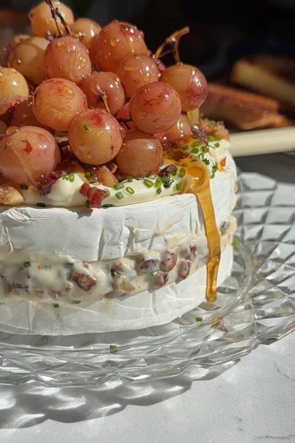 Roasted grape brie cake on a glass stand with honey dripping down the side and the pancetta walnut filling visible in the middle layer