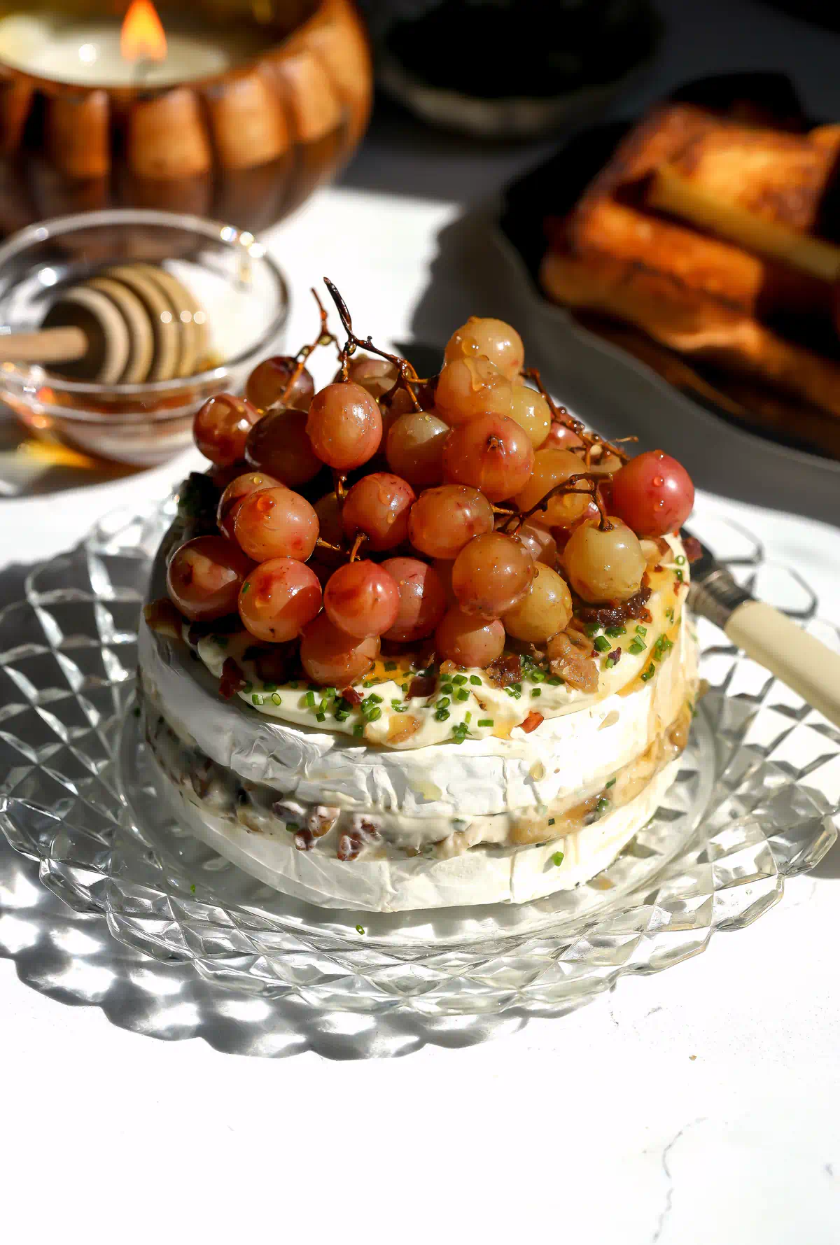 Roasted grape brie cake topped with a cluster of grapes still on the vine, pancetta, walnuts and chives, with a cheese knife on the plate