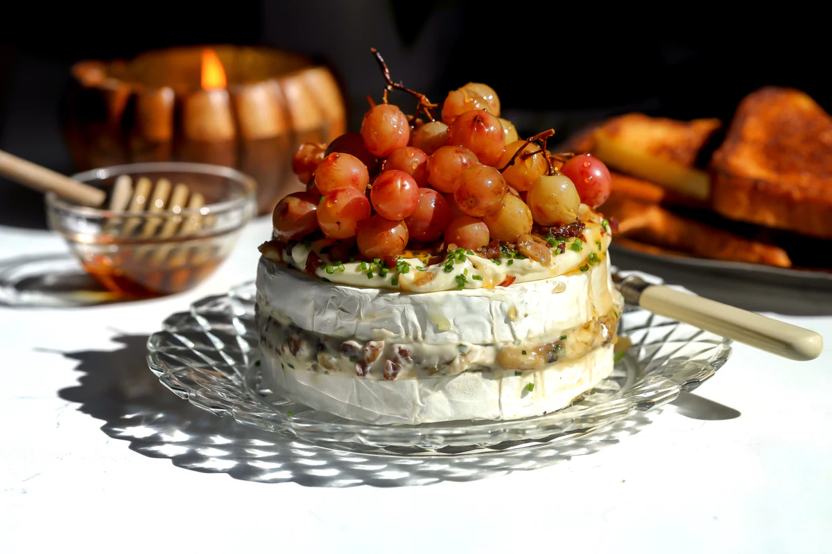 Roasted grape brie cake on a glass plate next to a bowl of honey and a platter of toasted bread