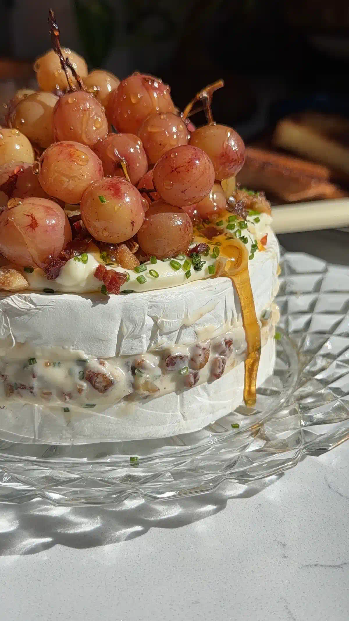 Roasted grape brie cake on a glass stand with honey dripping down the side and the pancetta walnut filling visible in the middle layer