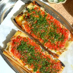 Overhead view of two tomato toasts topped with chives and black pepper, next to a bowl of chopped chives and a dish of garlic confit in olive oil.