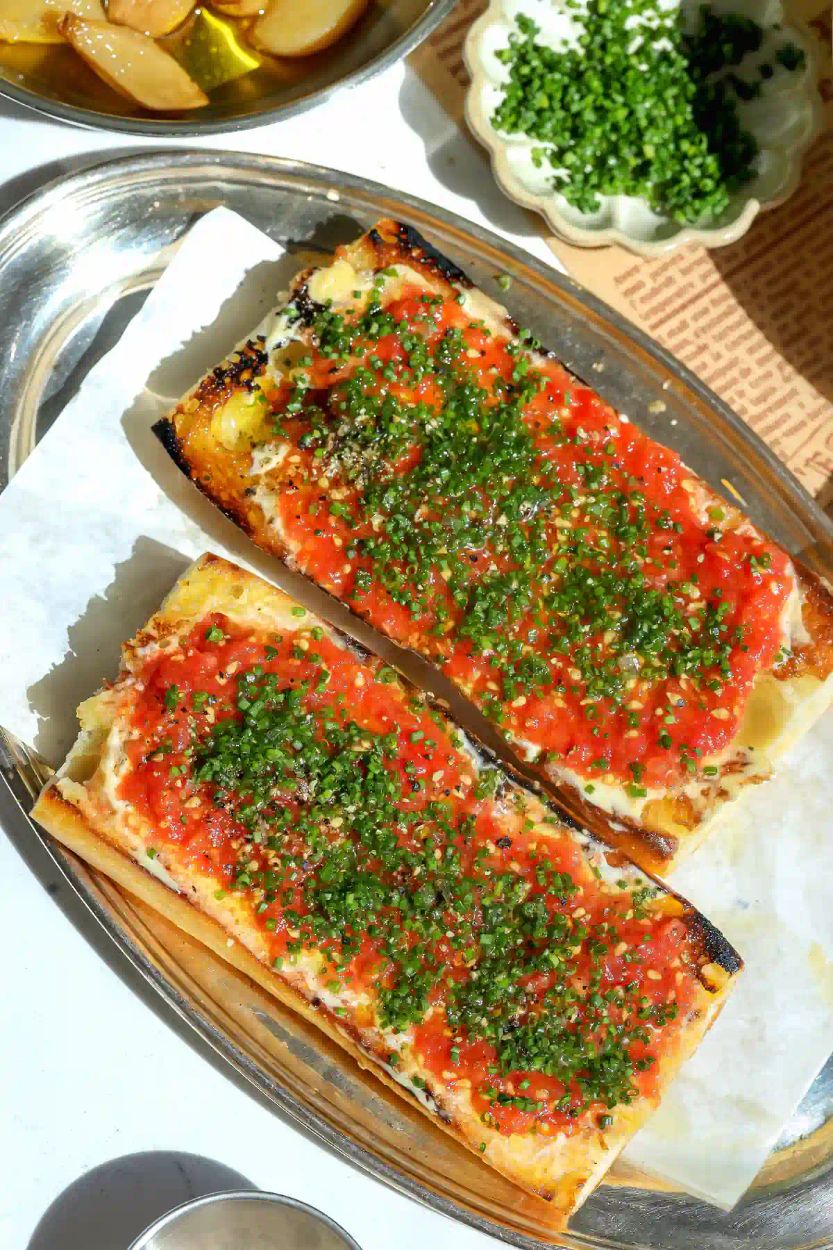 Overhead view of two tomato toasts topped with chives and black pepper, next to a bowl of chopped chives and a dish of garlic confit in olive oil.