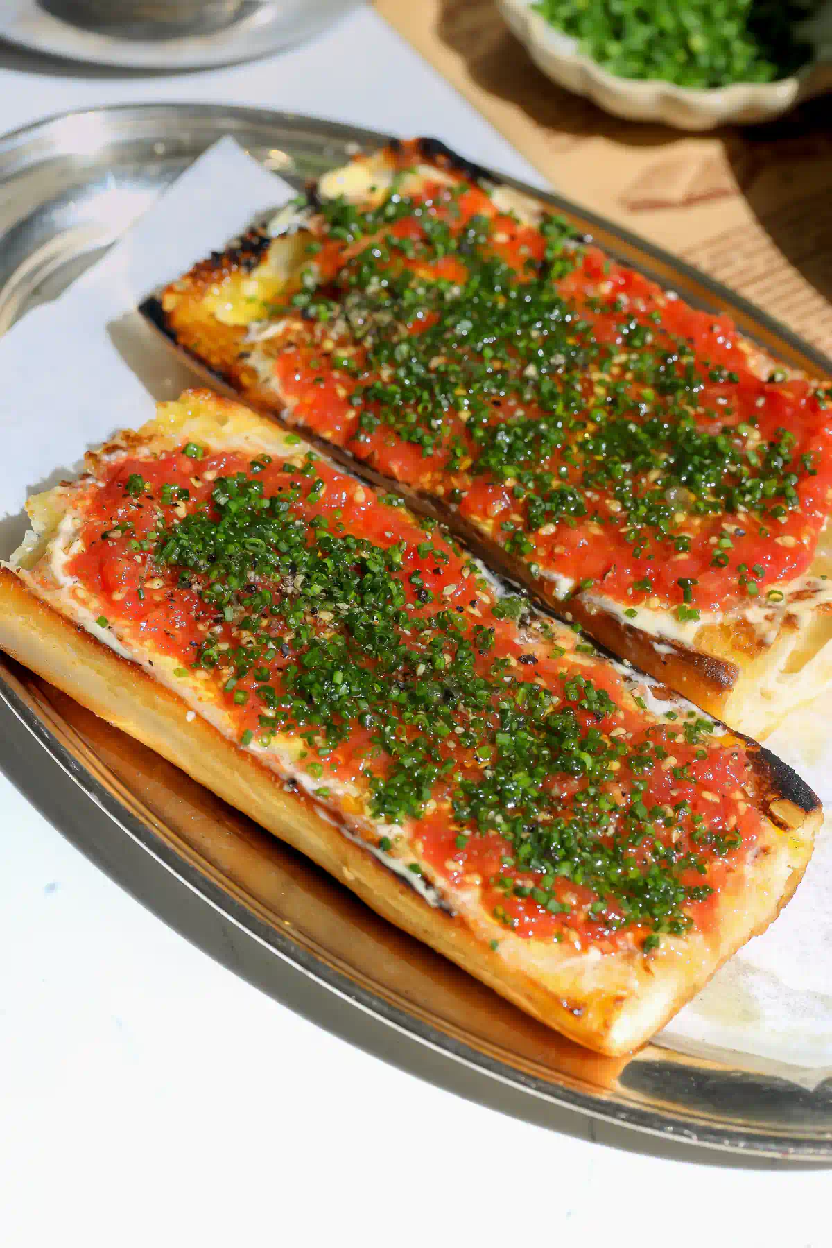 Two halves of a toasted baguette topped with grated tomato, chives, and black pepper on a metal tray lined with parchment.