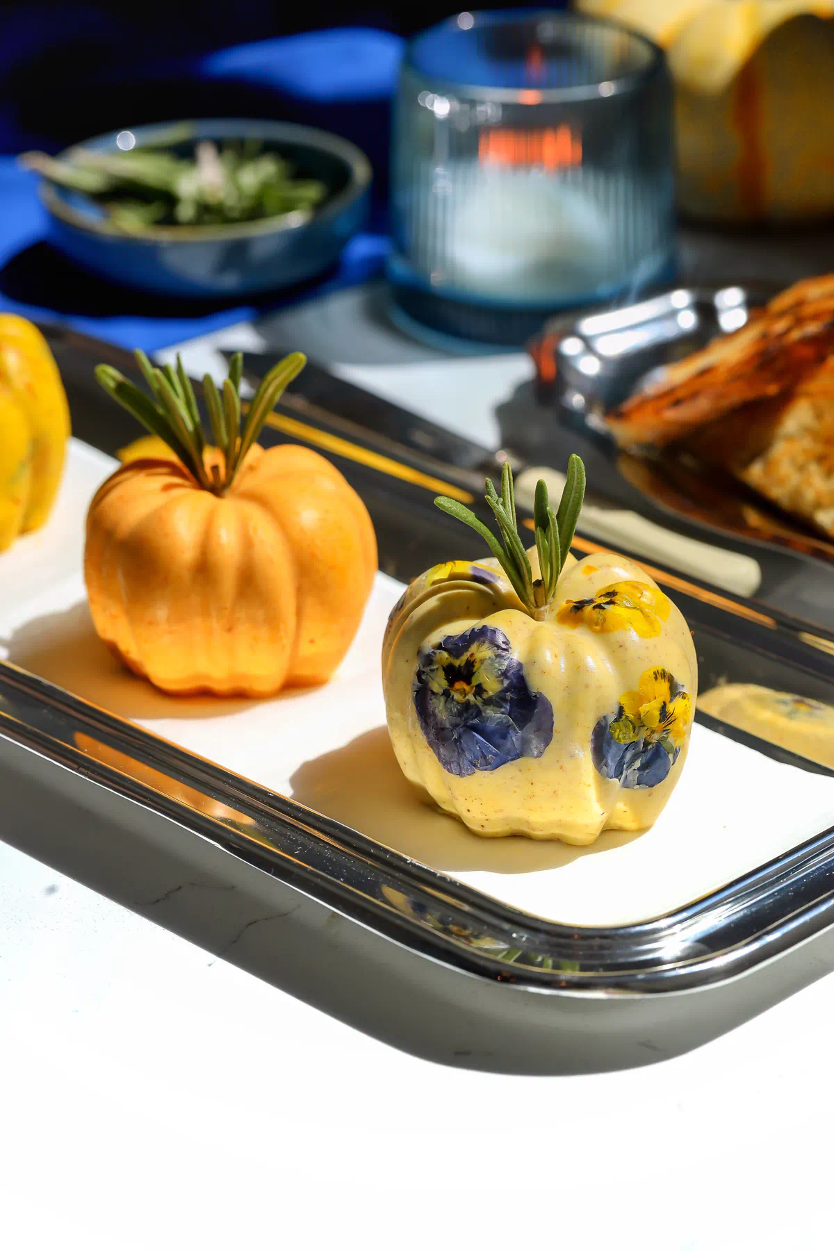 Butter pumpkins with rosemary stems on a silver tray, with purple and yellow edible flowers pressed into the maple pecan butter.