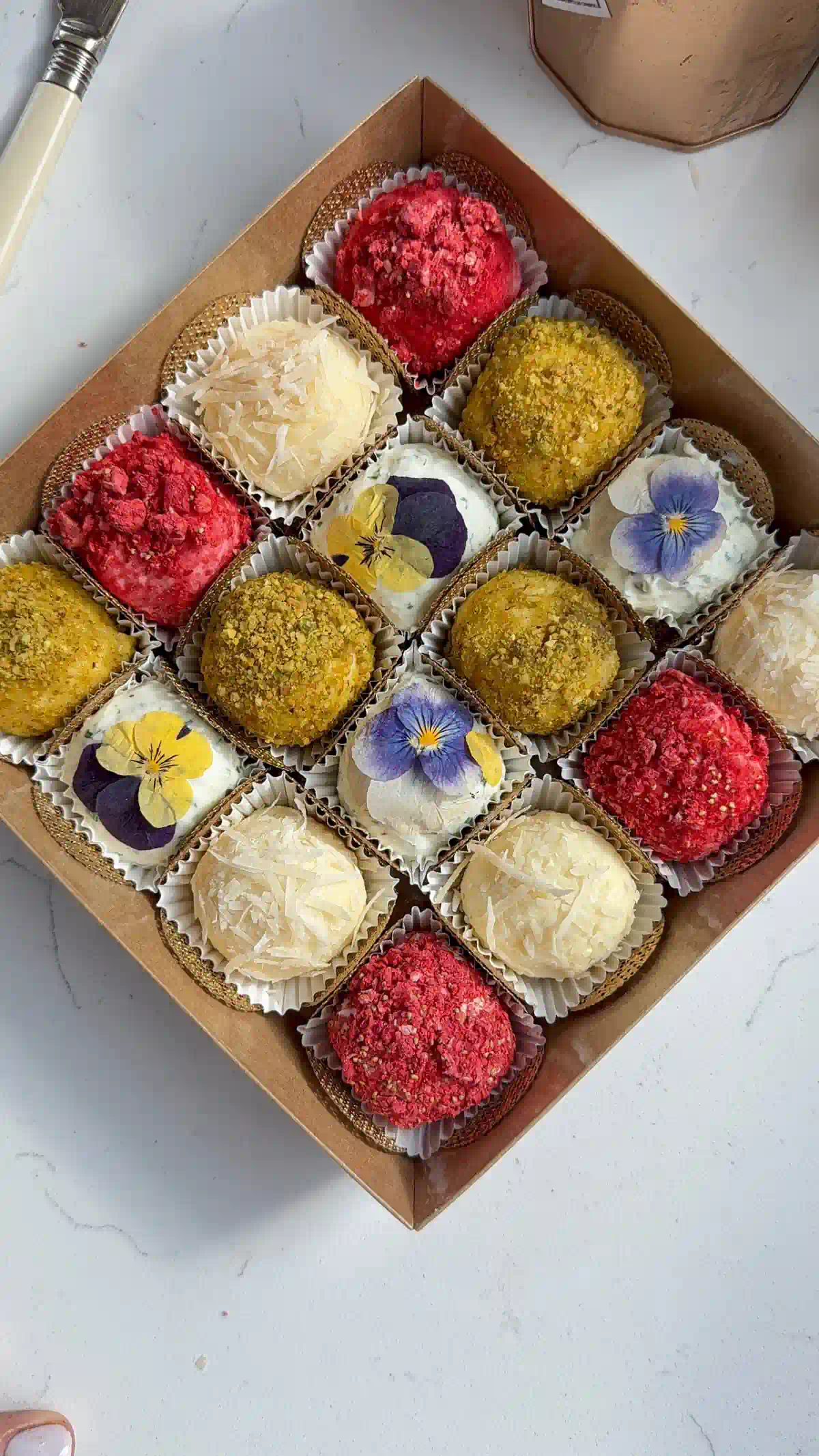 Cheese truffles coated in pistachios, raspberries, and parmesan or topped with edible flowers, arranged in a gift box.