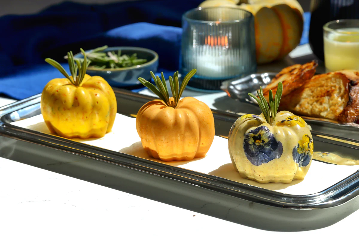 Three butter pumpkins topped with rosemary sprigs on a silver platter beside toasted bread and a lit candle.