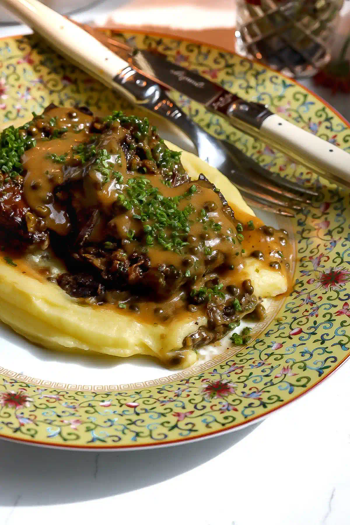 Close-up of browned short ribs with whole green peppercorns in cream sauce, chopped chives, and smooth potato purée.