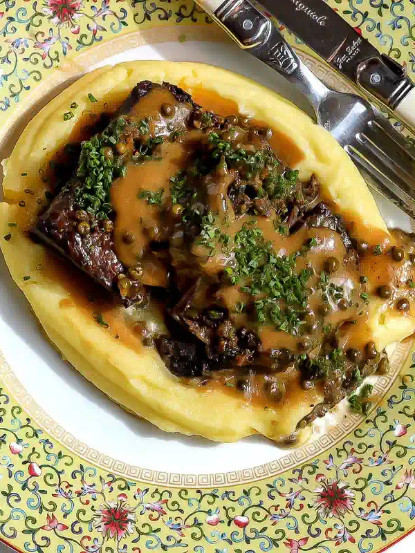 Overhead view of braised short ribs over potato purée, topped with green peppercorn cream sauce and chopped chives.