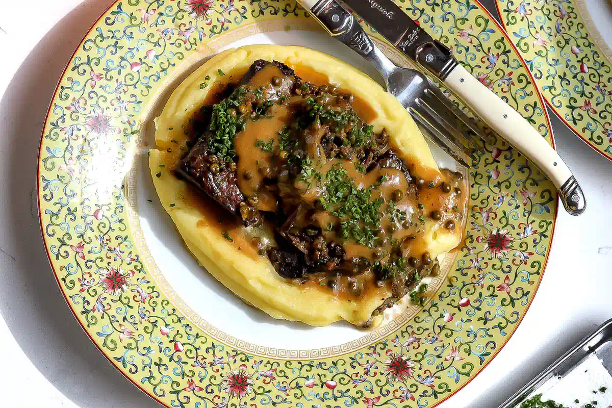 Overhead view of braised short ribs over potato purée, topped with green peppercorn cream sauce and chopped chives.