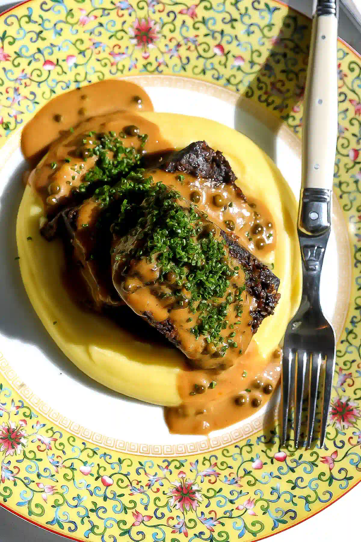 Peppercorn-crusted short ribs covered in cream sauce and chives, nestled in potato purée on a yellow floral plate.