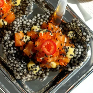 Close-up of salmon tartare in a puffed squid ink tapioca crisp with miso ginger mayo, black sesame seeds, trout roe, and a ponzu pipette.