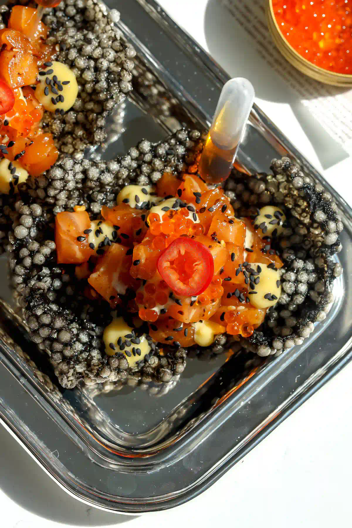 Close-up of salmon tartare in a puffed squid ink tapioca crisp with miso ginger mayo, black sesame seeds, trout roe, and a ponzu pipette.