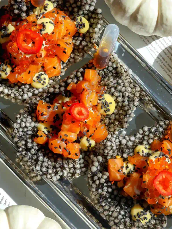 Overhead view of squid ink tapioca crisps filled with salmon tartare, miso ginger mayo, trout roe, and red chilies on a silver platter.