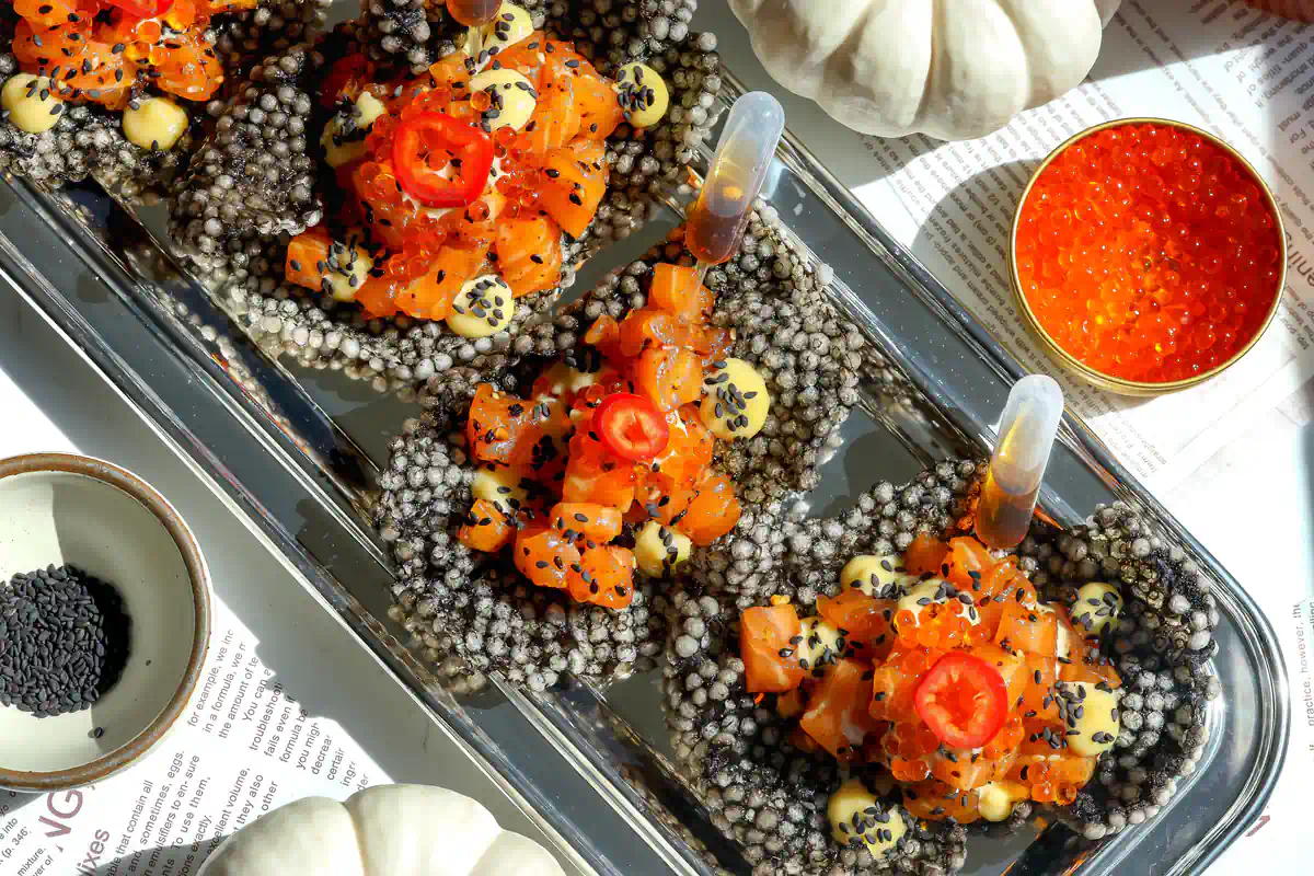 Overhead view of squid ink tapioca crisps filled with salmon tartare, miso ginger mayo, trout roe, and red chilies on a silver platter.