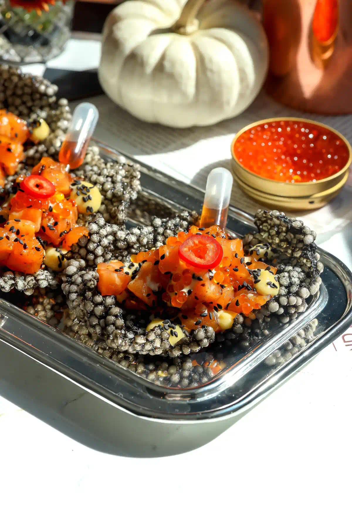 Angled view of squid ink tapioca crisps topped with diced salmon, trout roe, red chili slices, and miso ginger mayo dotted with black sesame seeds.
