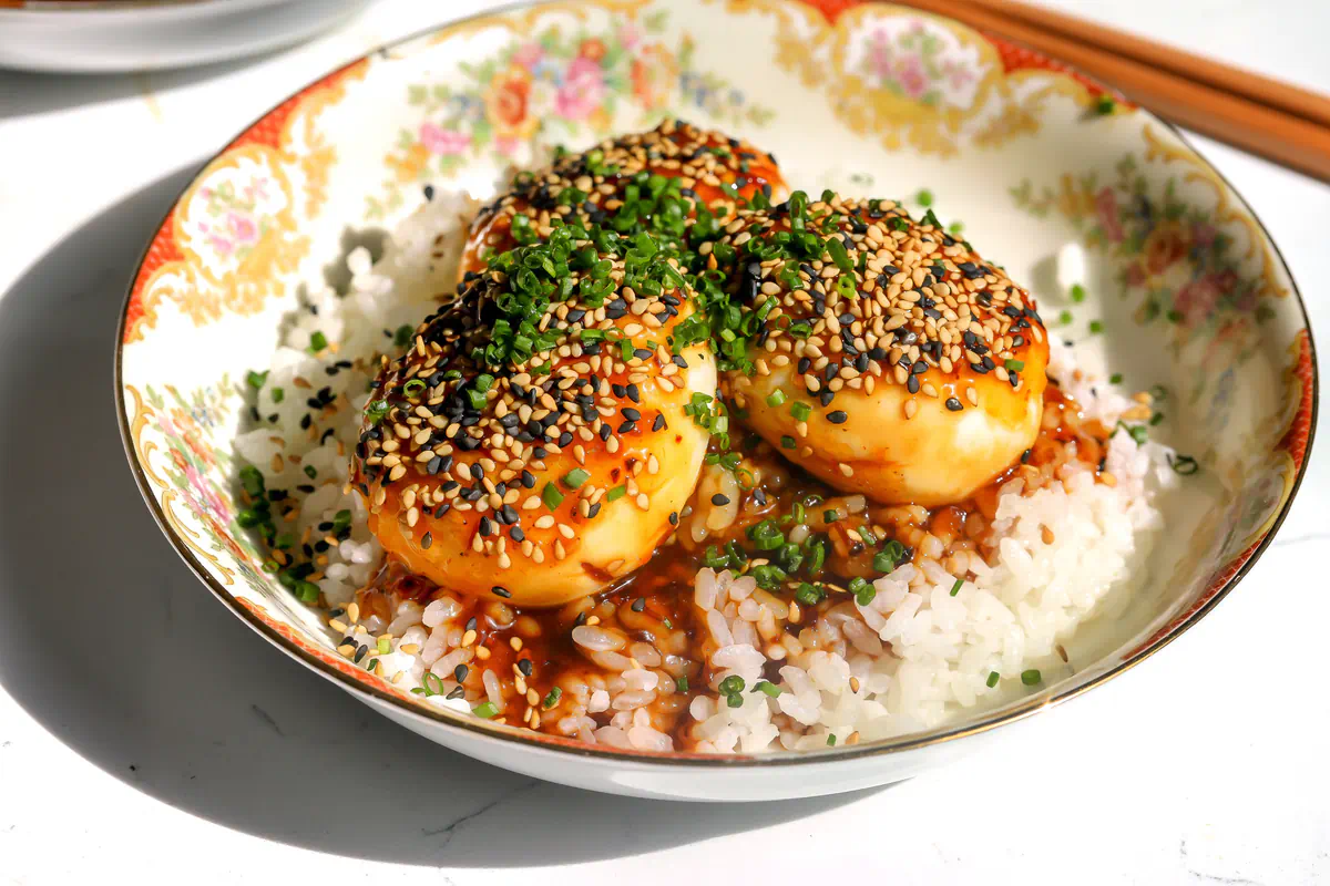 Three tiger skin eggs with sauce, sesame seeds, and chives over white rice in a floral bowl, with chopsticks resting across the rim.